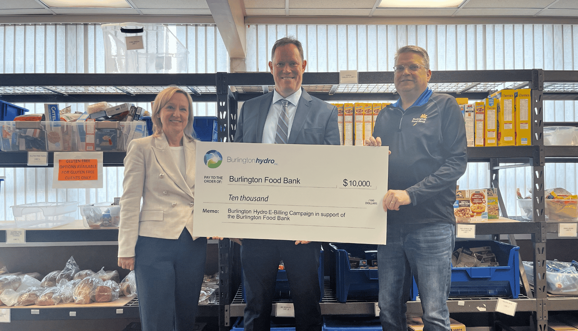 Three people pose in a food bank, holding a large £10,000 donation cheque from Burlington Hydro.