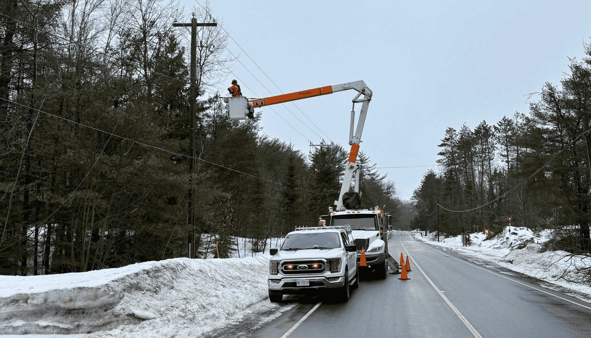 A utility worker in a cherry picker repairs power lines by the roadside on a snowy, overcast day.