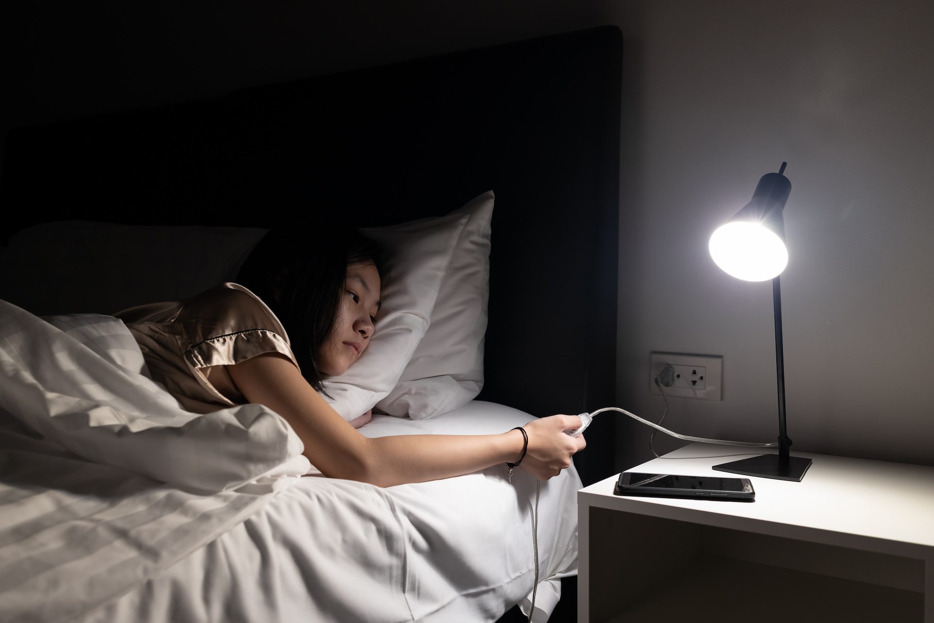 A person lying in bed at night reaches to plug a phone into a charger on a bedside table with a lamp.
