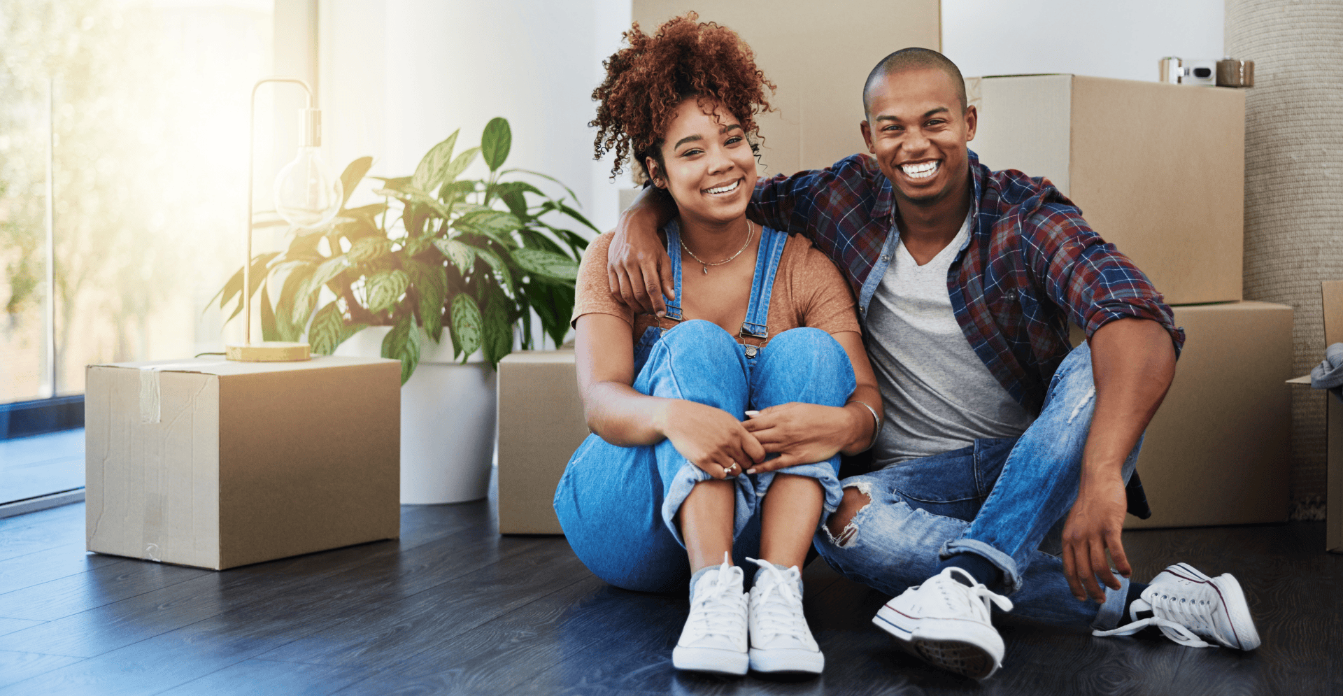 A couple sit on the floor of their home, smiling, surrounded by packing boxes and a potted plant.