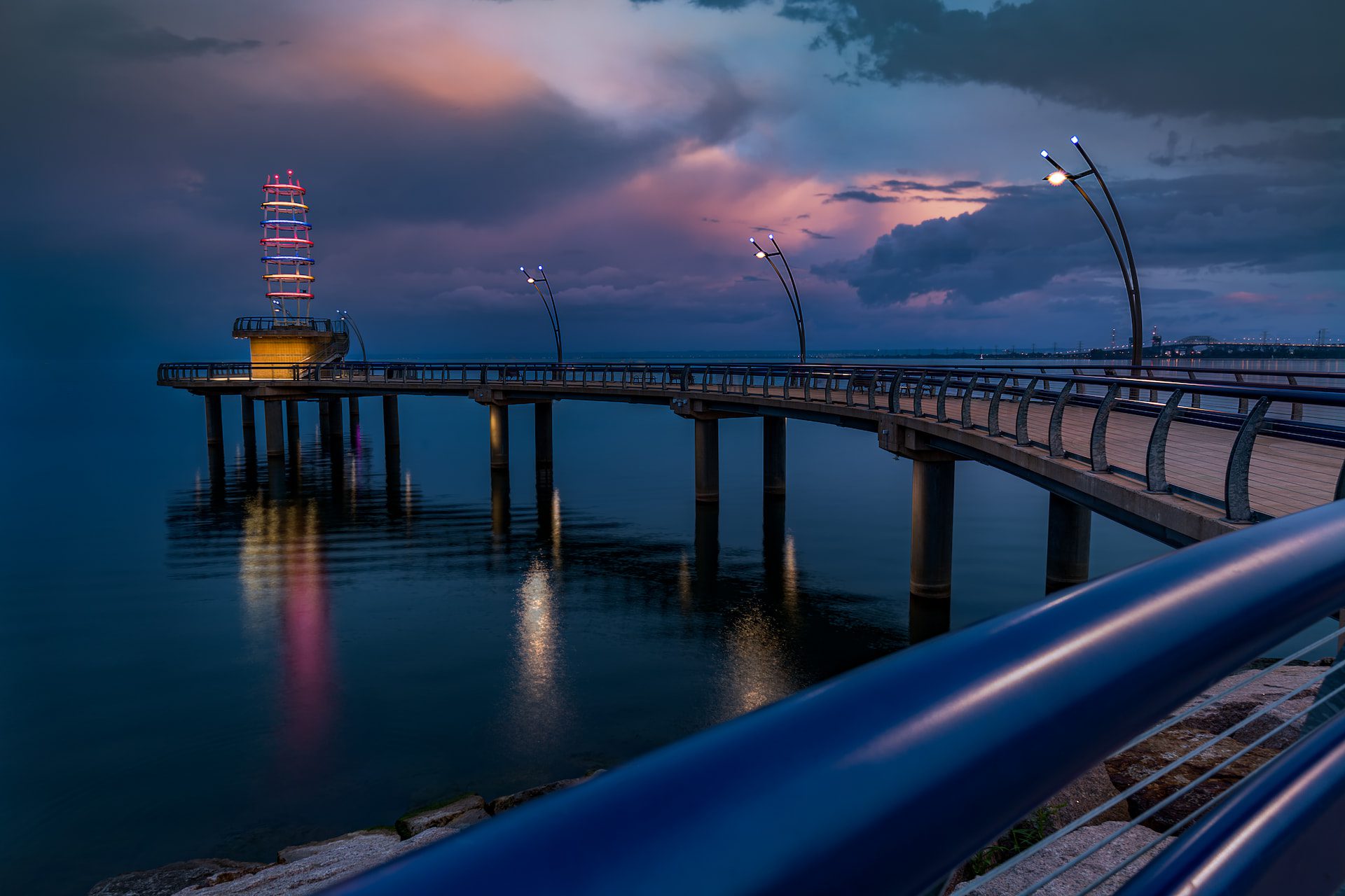 A long pier with railings extends over calm water at dusk, with a lit observation tower at the end under a cloudy sky.