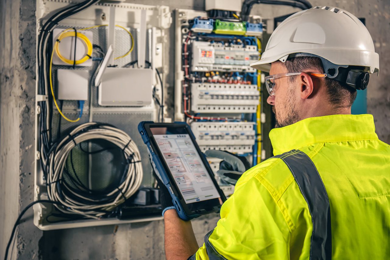 An engineer in safety gear inspects electrical panels and uses a tablet for data entry at an industrial site.