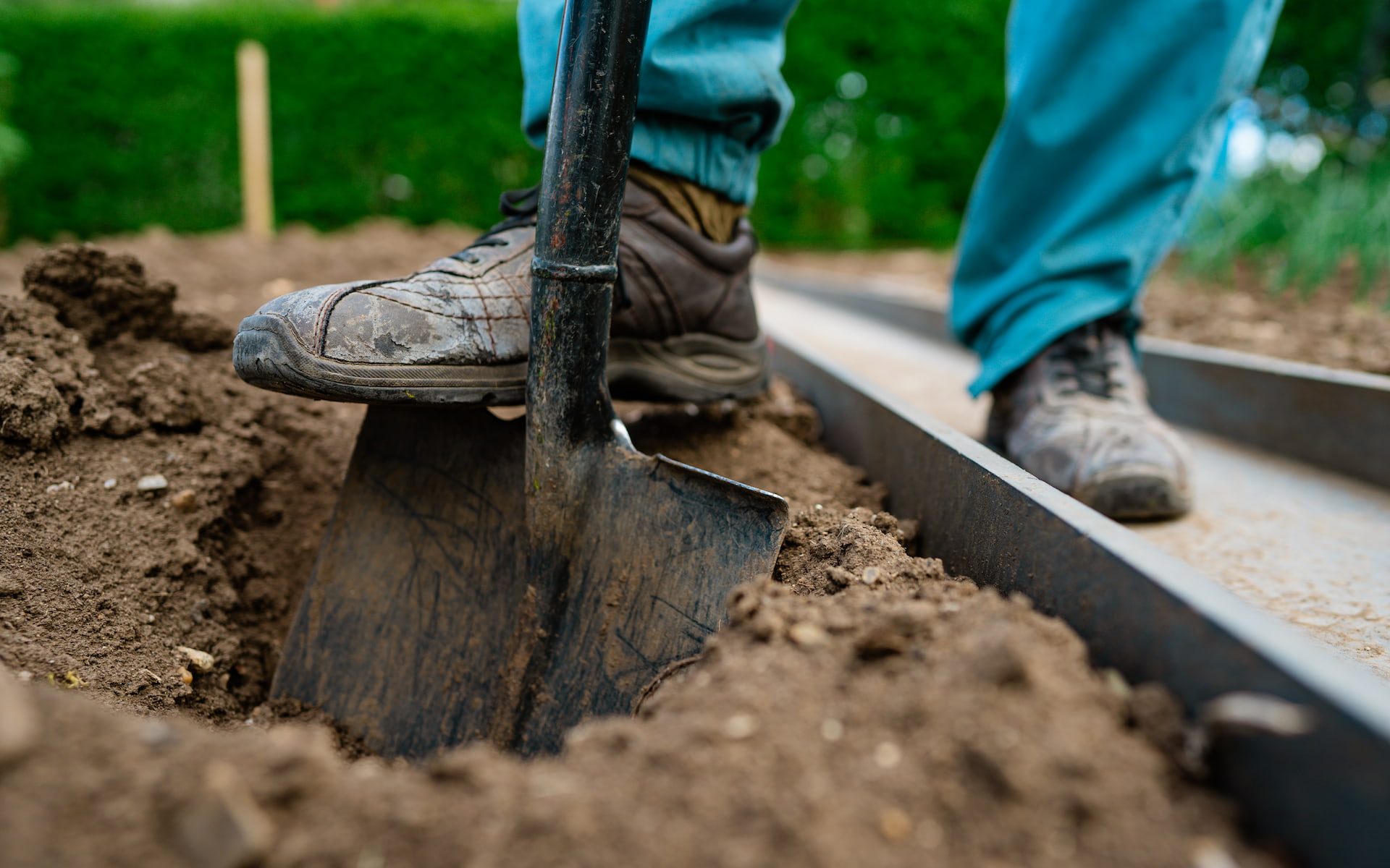 A person wearing work boots digs soil with a spade next to a metal edge outdoors.