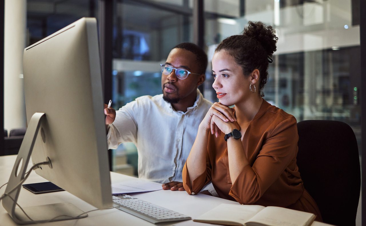 Two people sit at a desk working together on a computer in a modern office setting.
