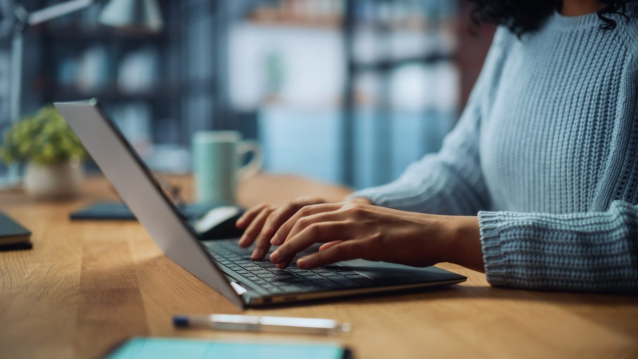 Person in a blue jumper types on a laptop at a wooden desk with a mug and plant in the background.
