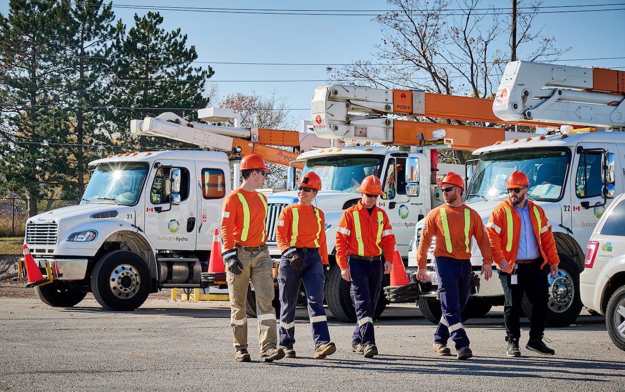 Five utility workers in orange safety gear walk in front of service lorries in a car park on a clear day.