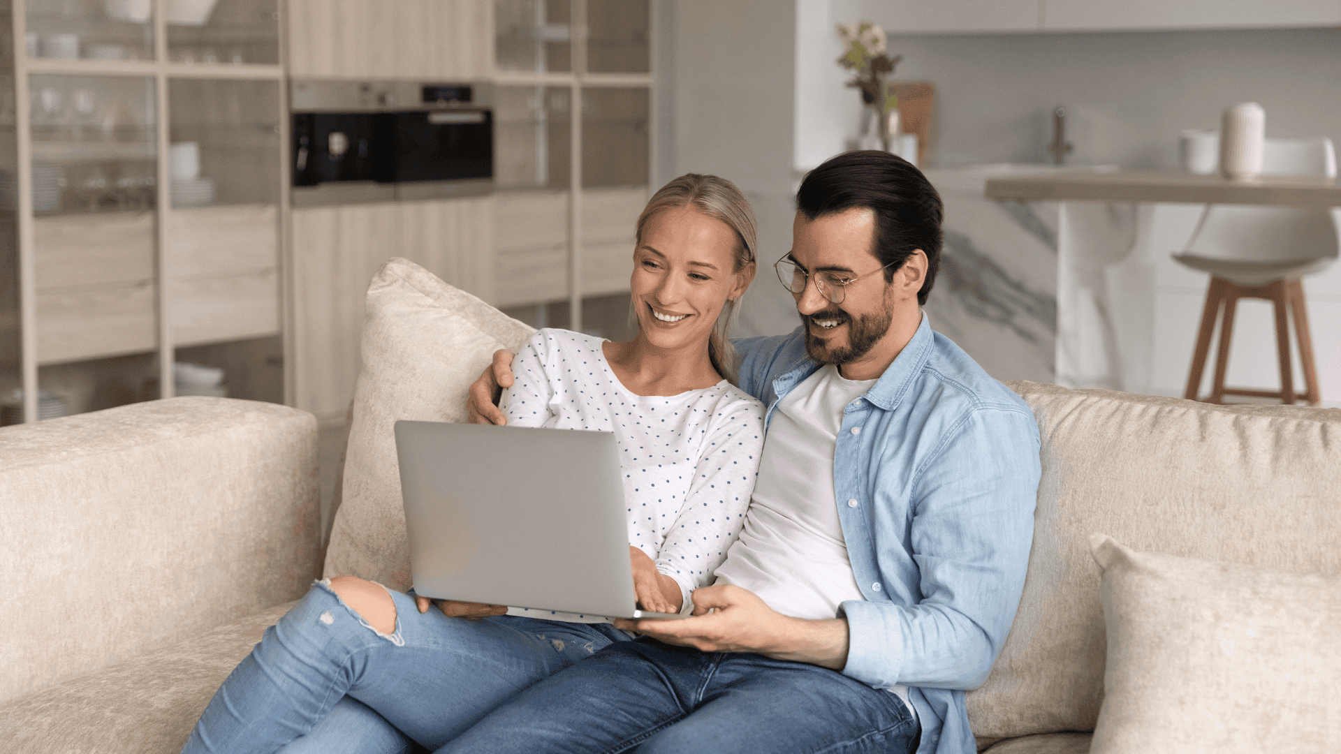 A couple sit together on a sofa, smiling while looking at a laptop screen in a modern lounge.