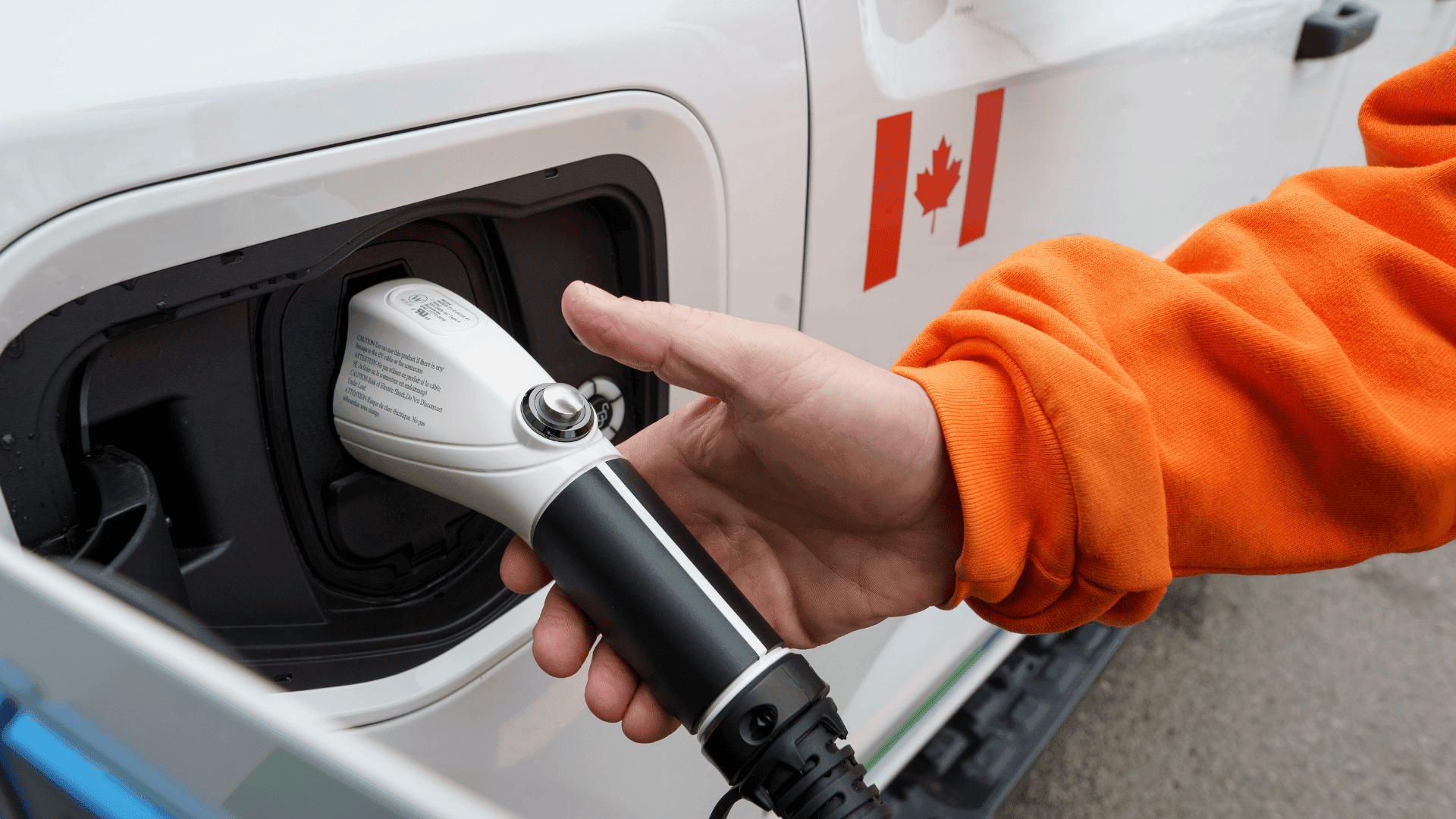 A person plugs an electric vehicle charging cable into a white vehicle with a Canadian flag sticker.