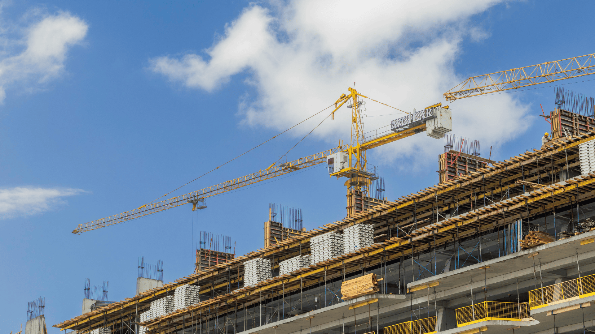 A yellow tower crane operates above a multi-storey concrete building under construction against a blue sky with clouds.