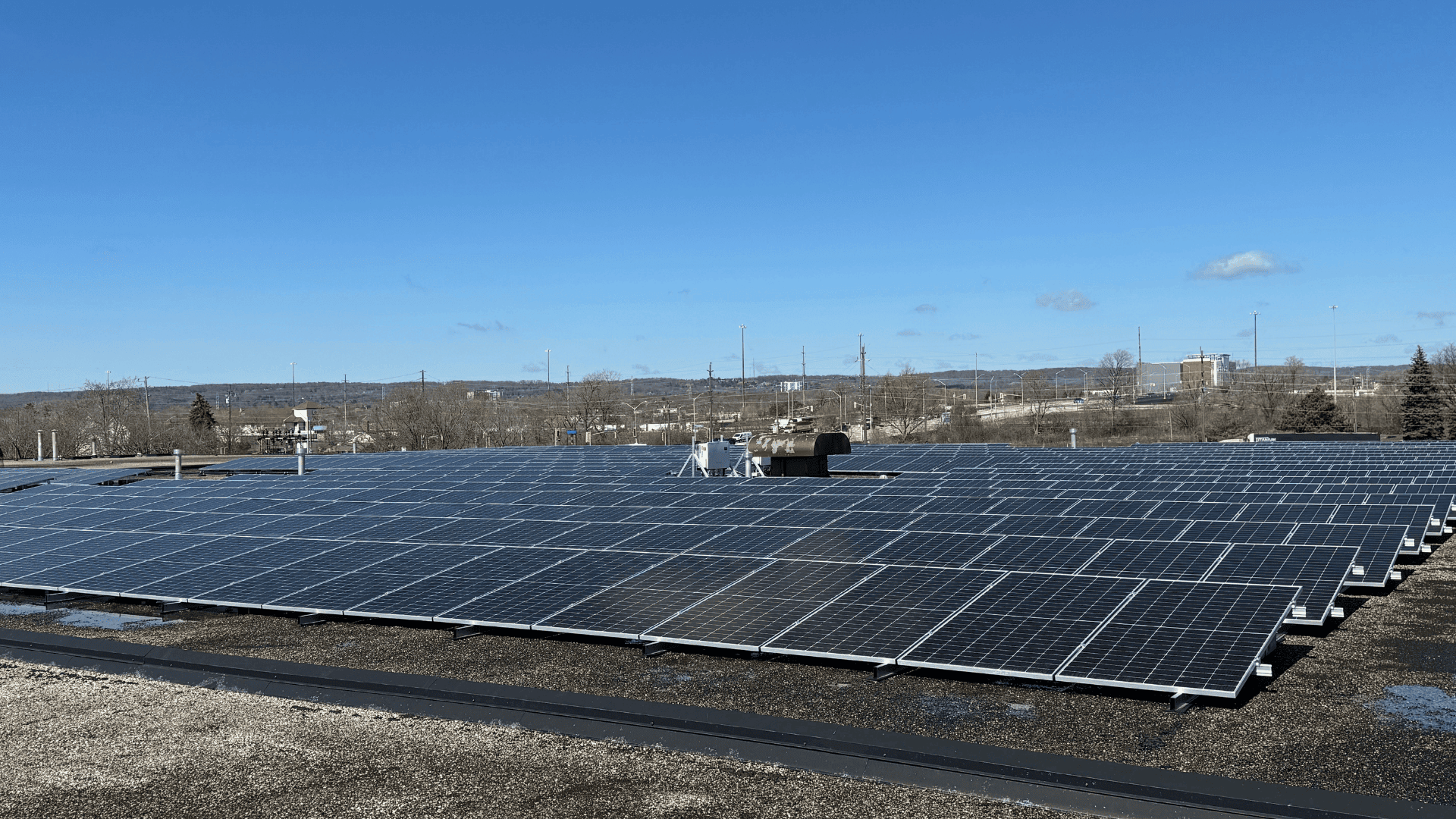 Rows of solar panels installed on a flat rooftop under a clear blue sky with distant buildings and trees in the background.