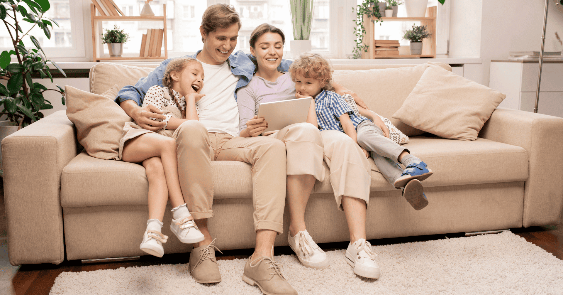 A family of four sits on a sofa, smiling and looking at a tablet together in a bright lounge.