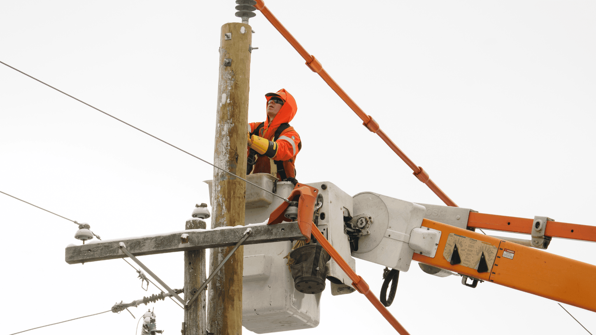 Utility worker in a cherry picker repairs power lines on a wooden telegraph pole against an overcast sky.