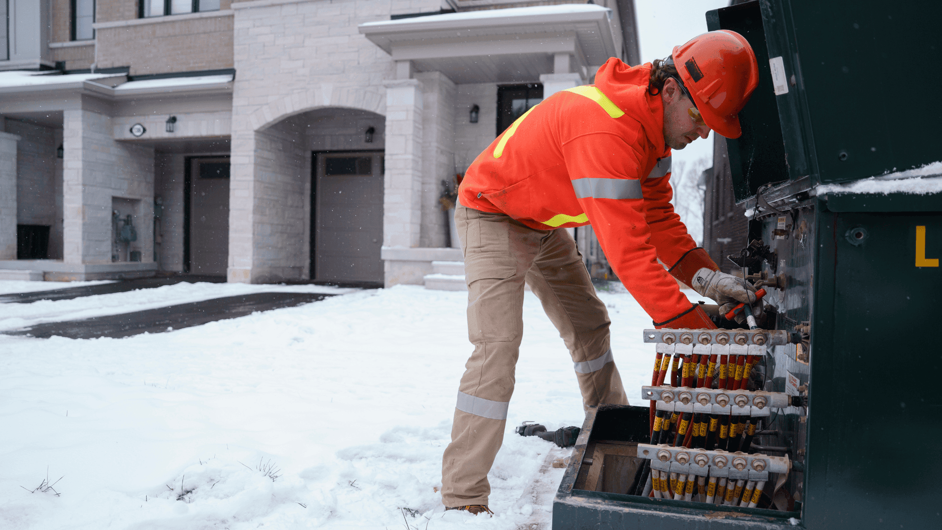 A utility worker in safety gear repairs electrical equipment outdoors in a snowy residential area.