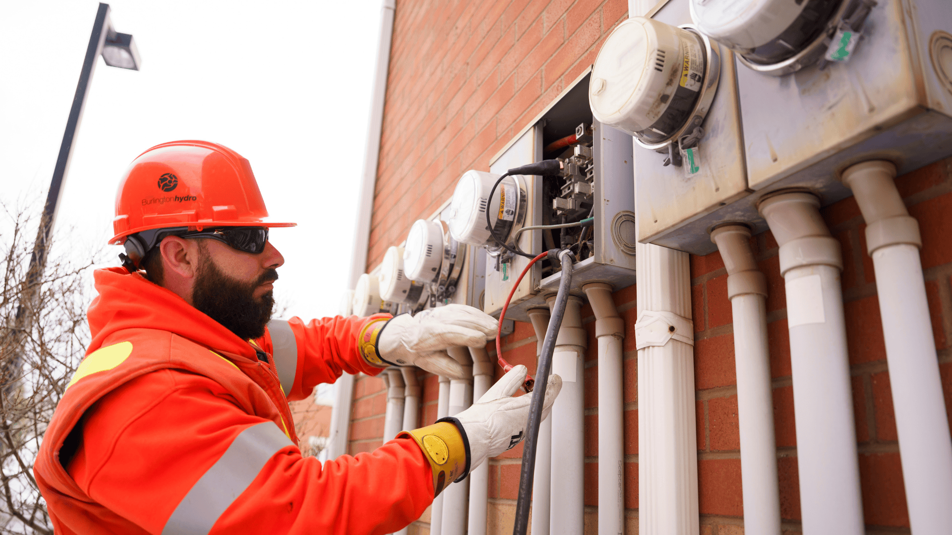 Utility worker in orange safety gear servicing electrical meters mounted on a brick wall outside.