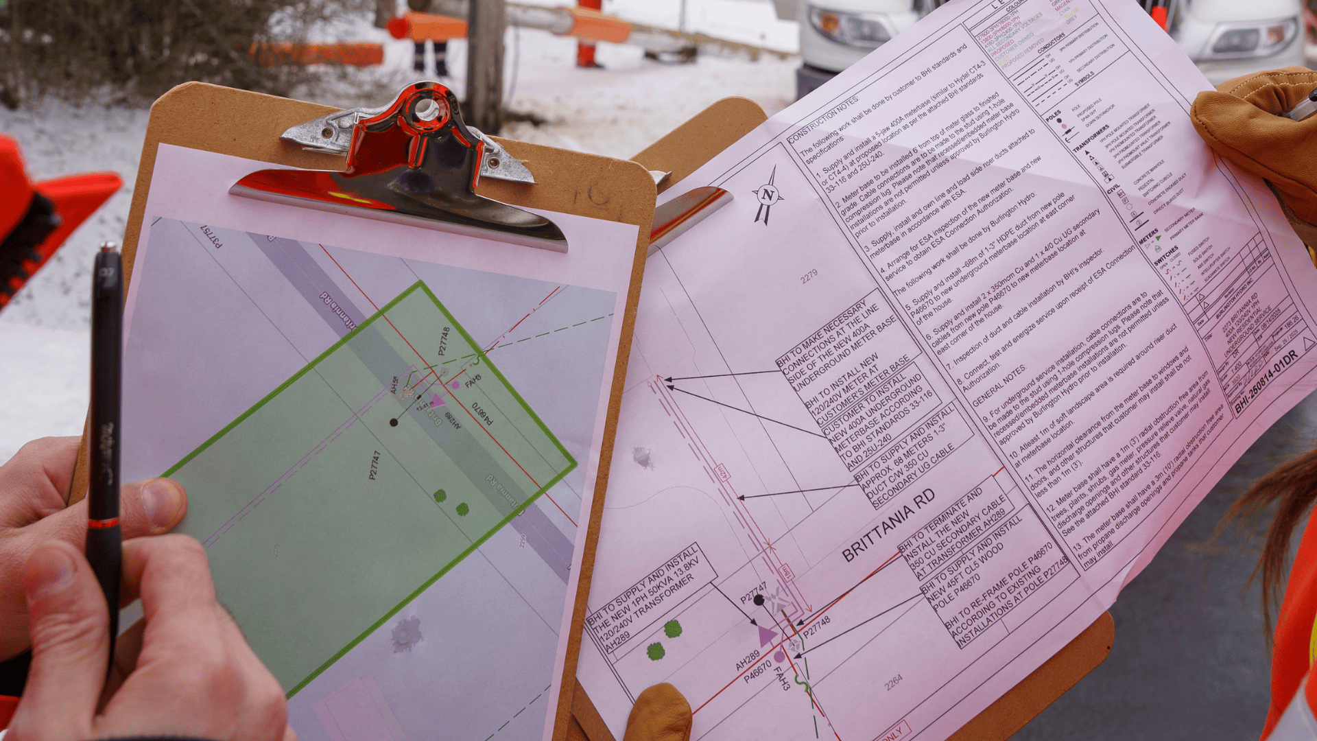 Two people hold clipboards with technical site maps and documents outdoors in a snowy construction area.