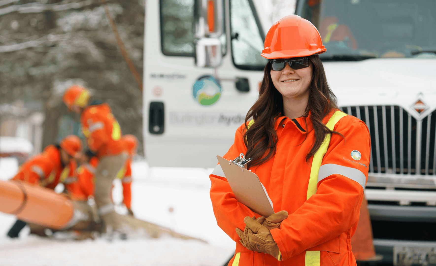 A worker in orange safety gear and helmet holds a clipboard in front of a utility lorry on a snowy day.