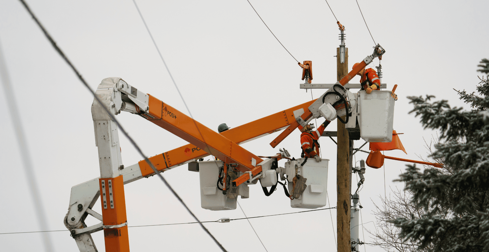 Utility workers in cherry pickers repair power lines on a wooden pole against a cloudy sky.