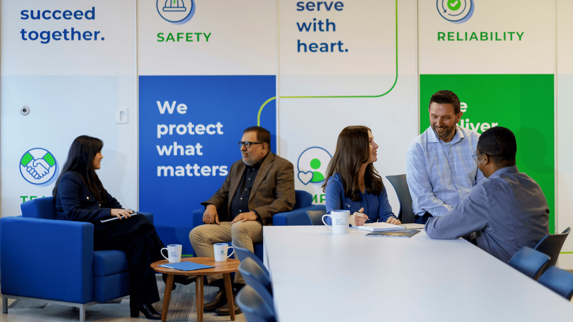 Six professionals have a discussion in a modern office with motivational and safety posters on the wall.