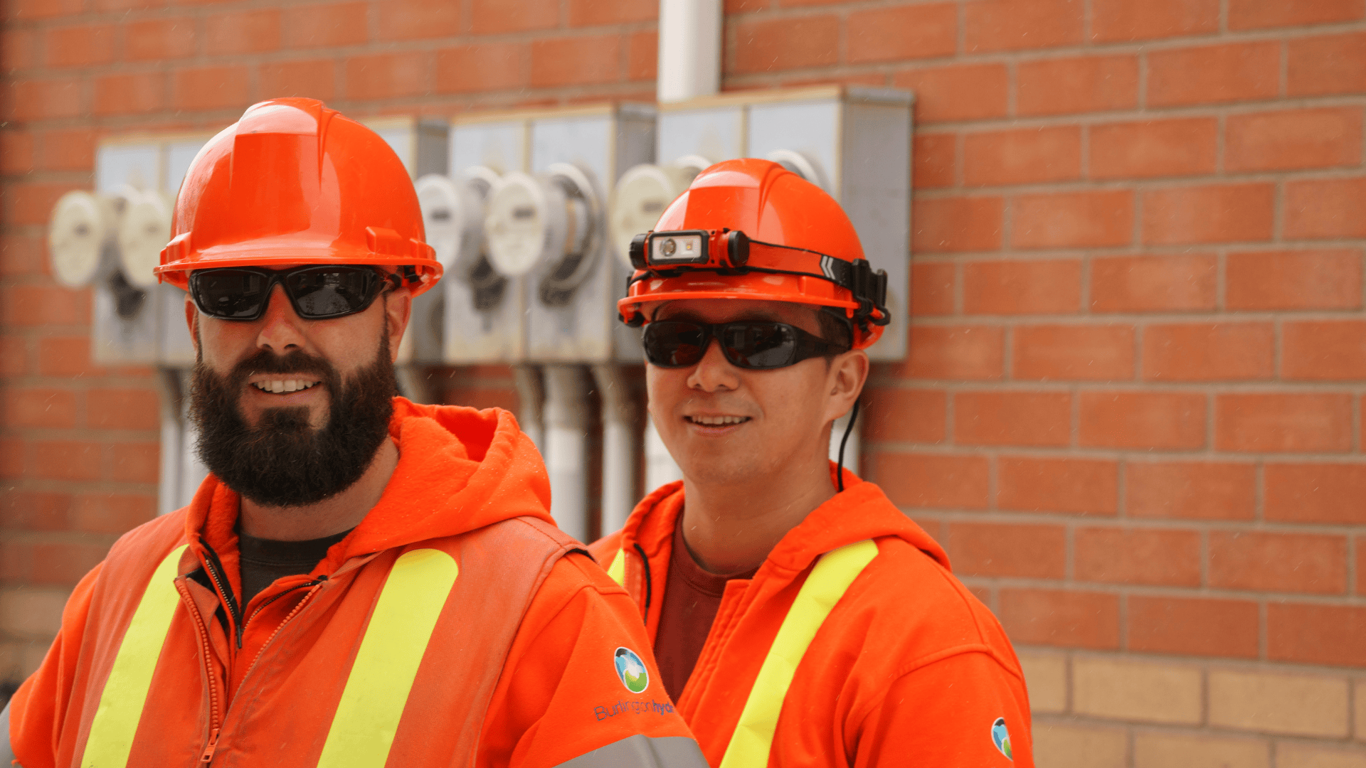 Two construction workers in orange safety gear and helmets stand in front of electricity meters on a brick wall.