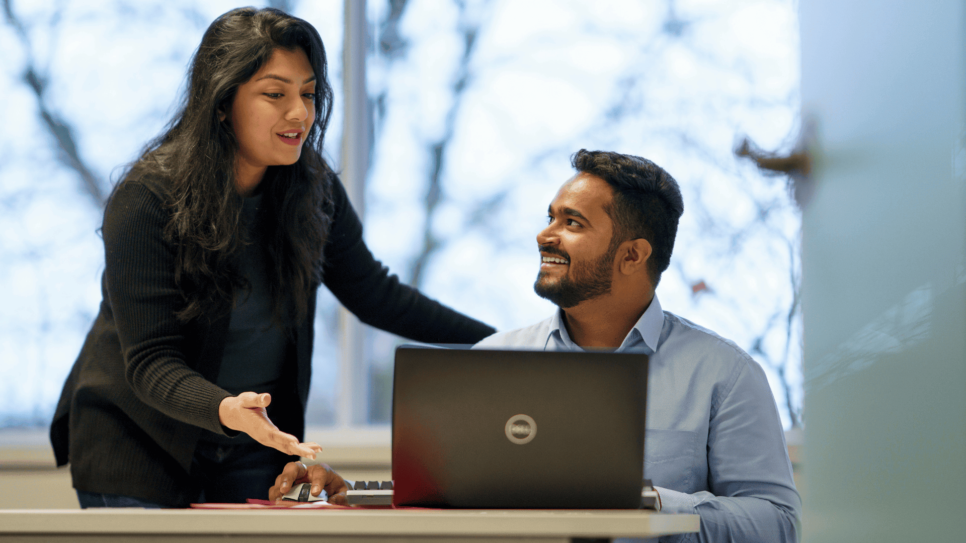 Woman gestures while talking to a man who is smiling and seated at a desk with a laptop in an office setting.
