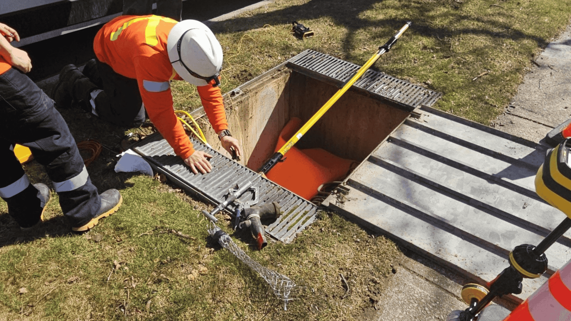 Two workers in safety gear inspect and work around an open manhole on a grassy area beside a pavement.