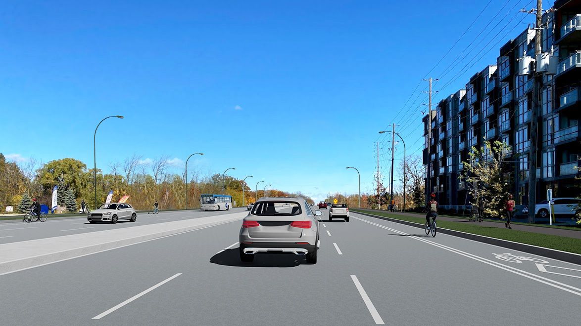 A wide city street with cars, a cyclist in a cycle lane, a bus, and pedestrians on a sunny day.