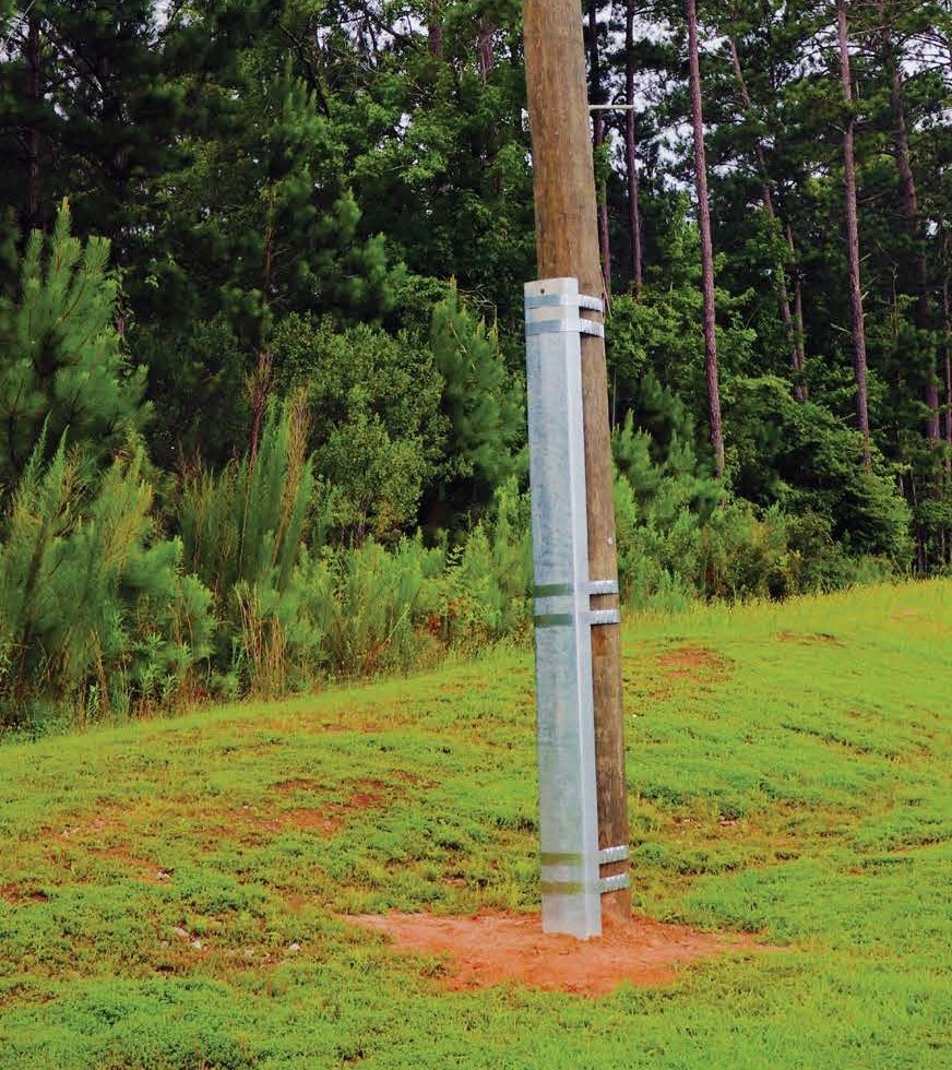 Wooden telegraph pole with power lines, reinforced at the base with a metal support, stands on grass near trees.