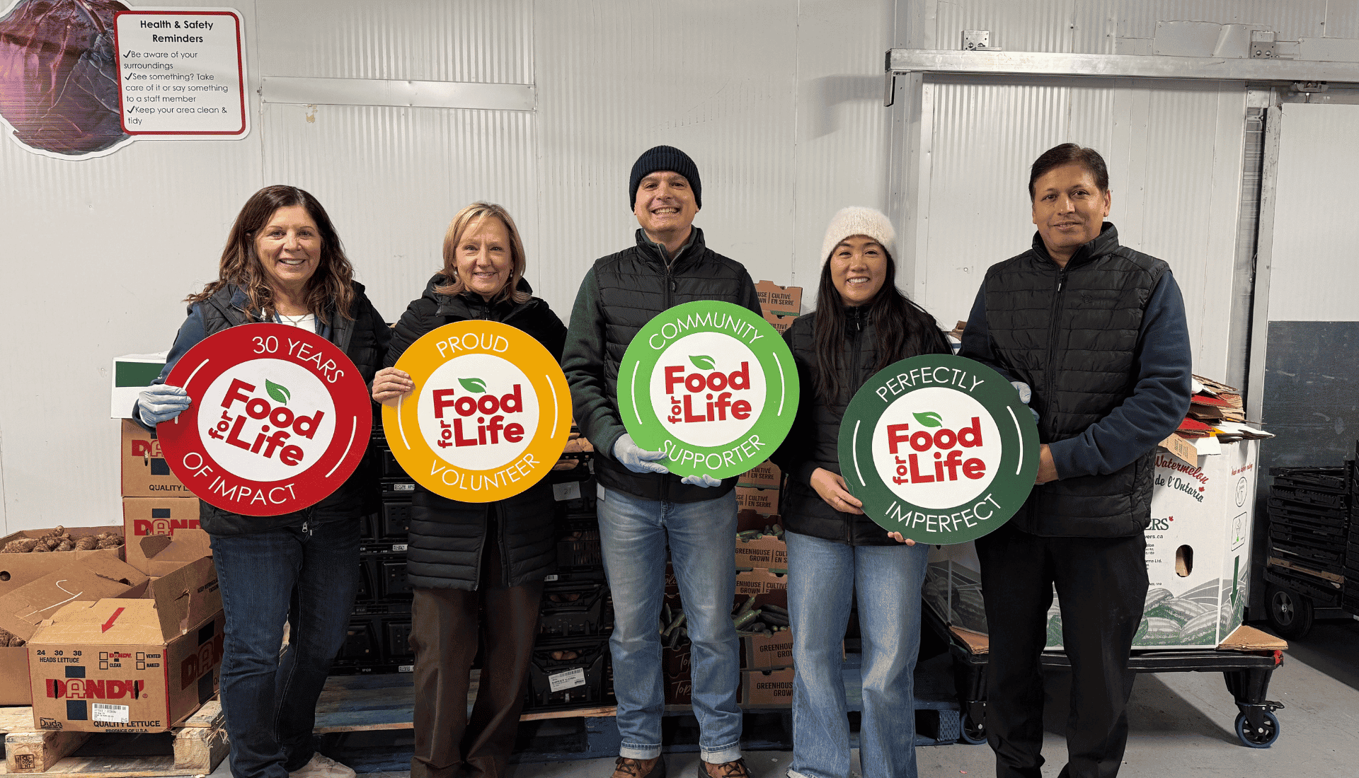 Five people stand indoors holding colourful Food for Life signs in front of boxes and crates at a food distribution centre.