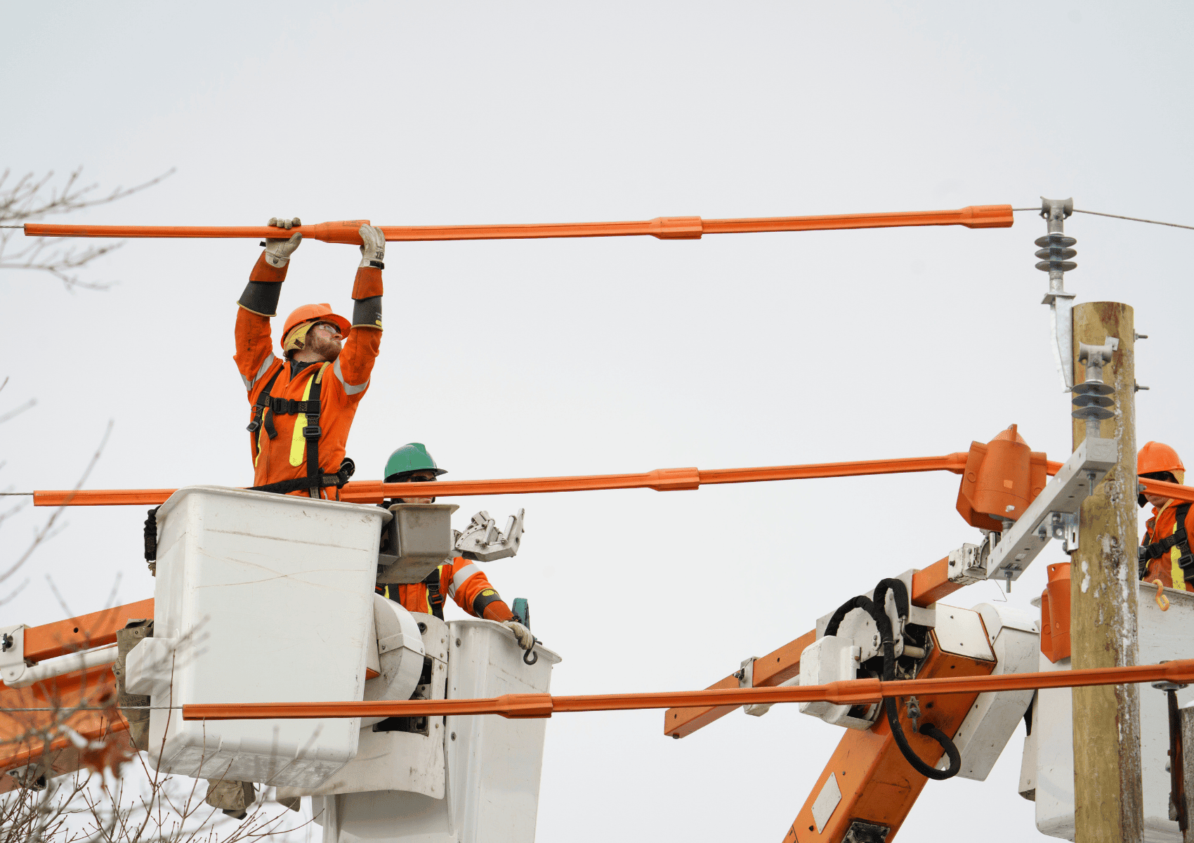 Two utility workers in cherry pickers use insulated poles to work on power lines and equipment.