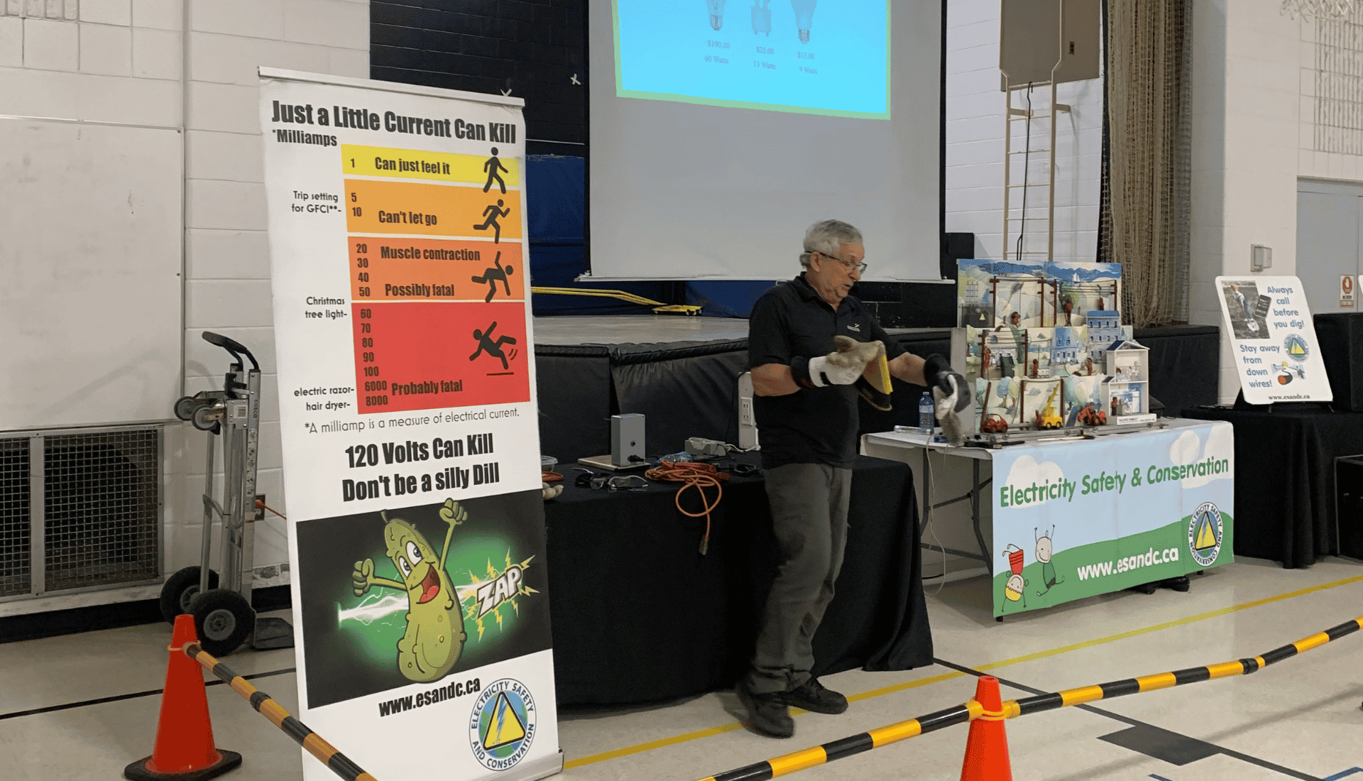 A man demonstrates electrical safety in a gymnasium with warning posters, props, and safety equipment on display tables.