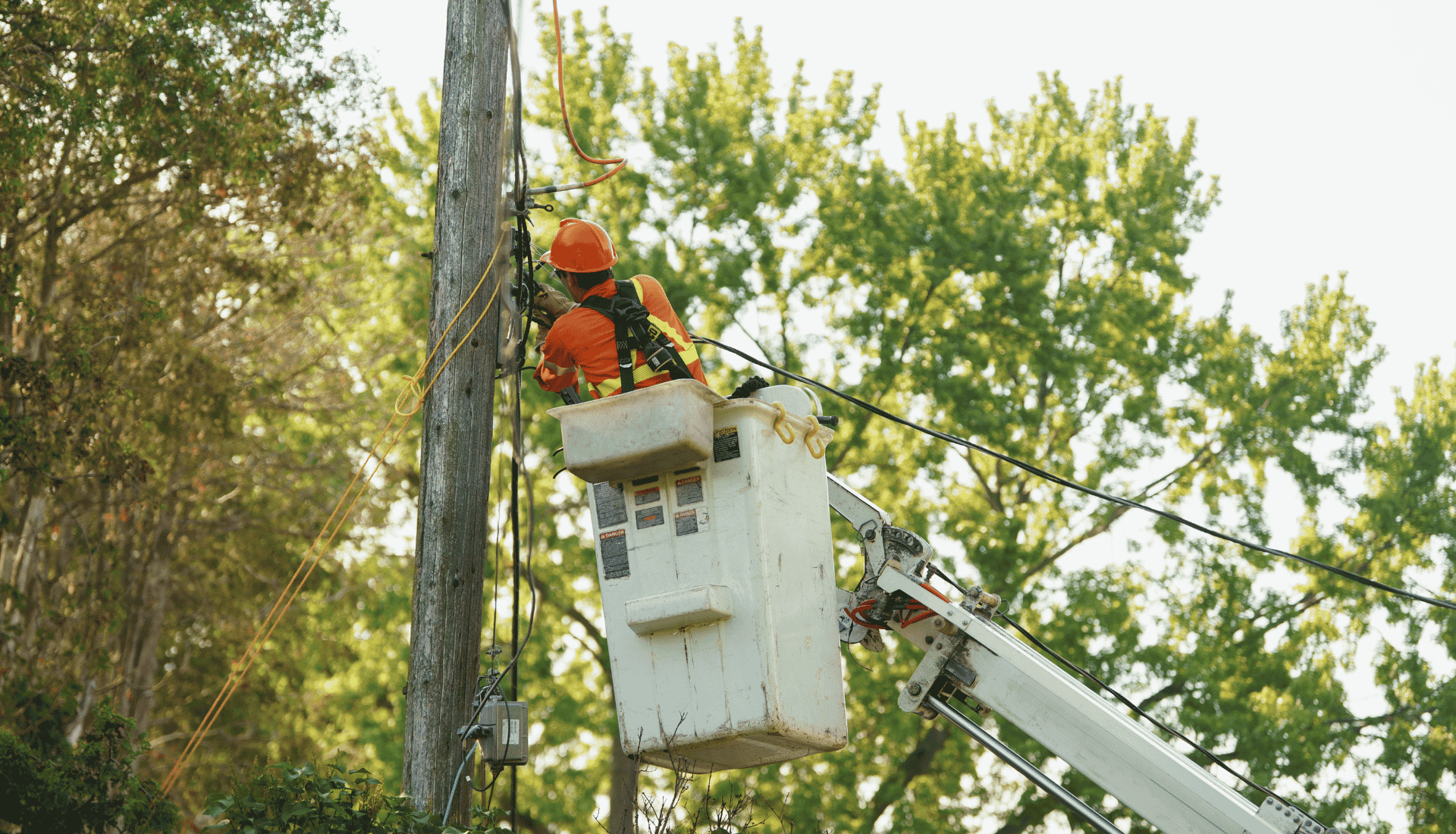 Utility worker in safety gear repairs wires on a telegraph pole from an elevated cherry picker amid trees.