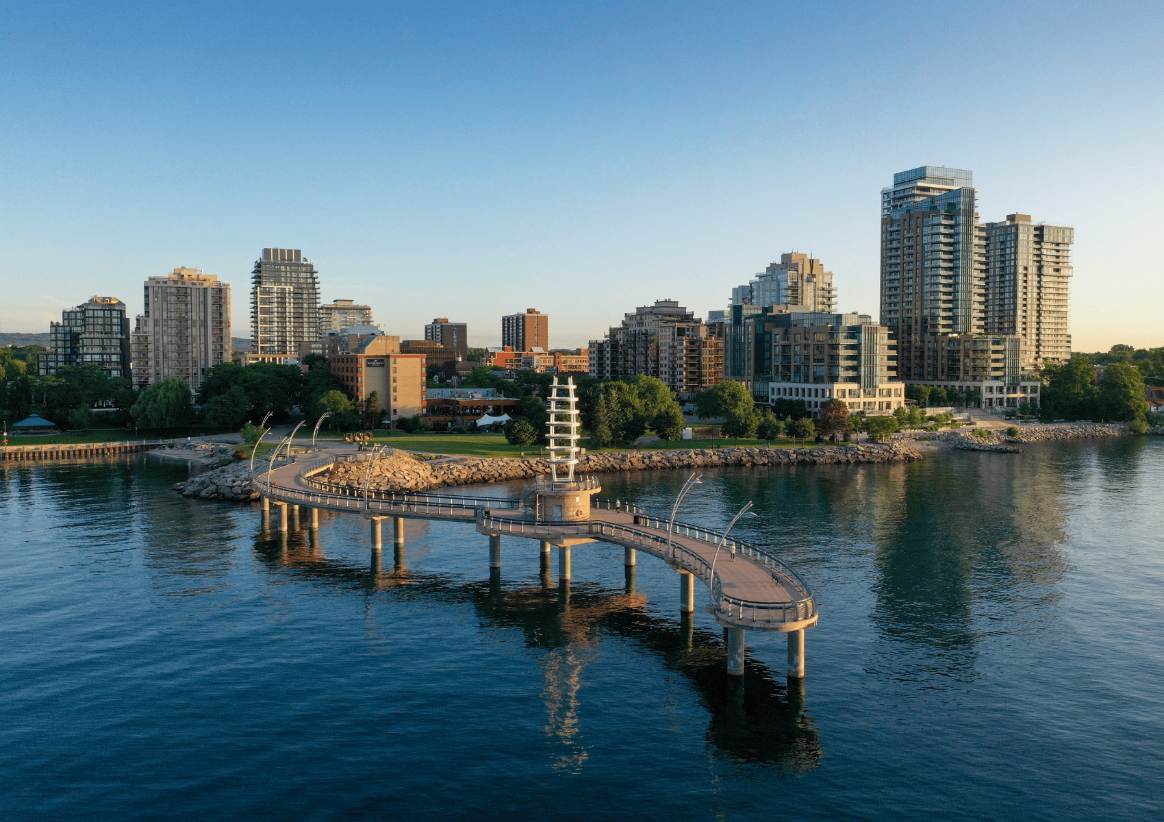 Curved pier with a tower extends over calm water, with city buildings and trees in the background at sunset.