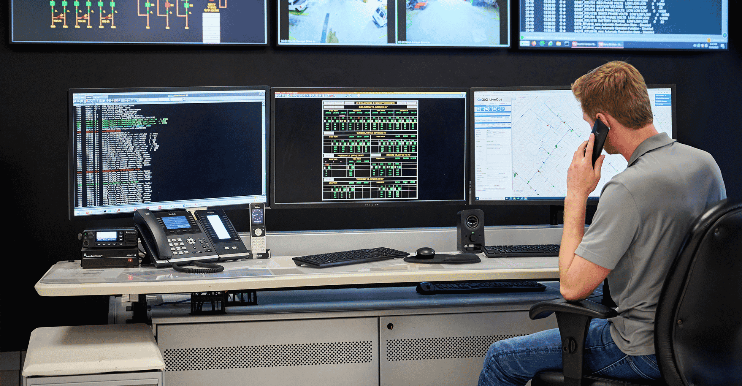 A person sits at a desk with multiple computer monitors, speaking on the phone in an office setting.