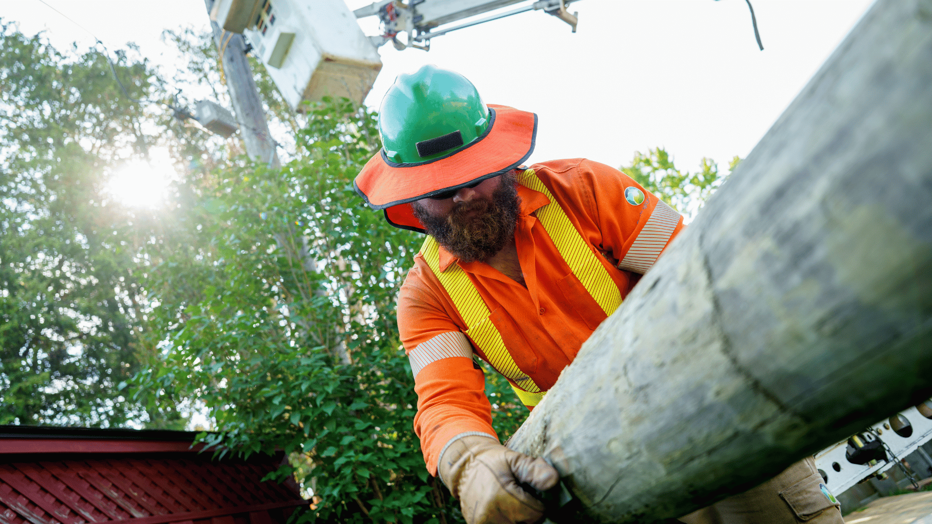 Utility worker in orange safety gear lifts a telegraph pole outdoors near trees with a utility lorry in the background.