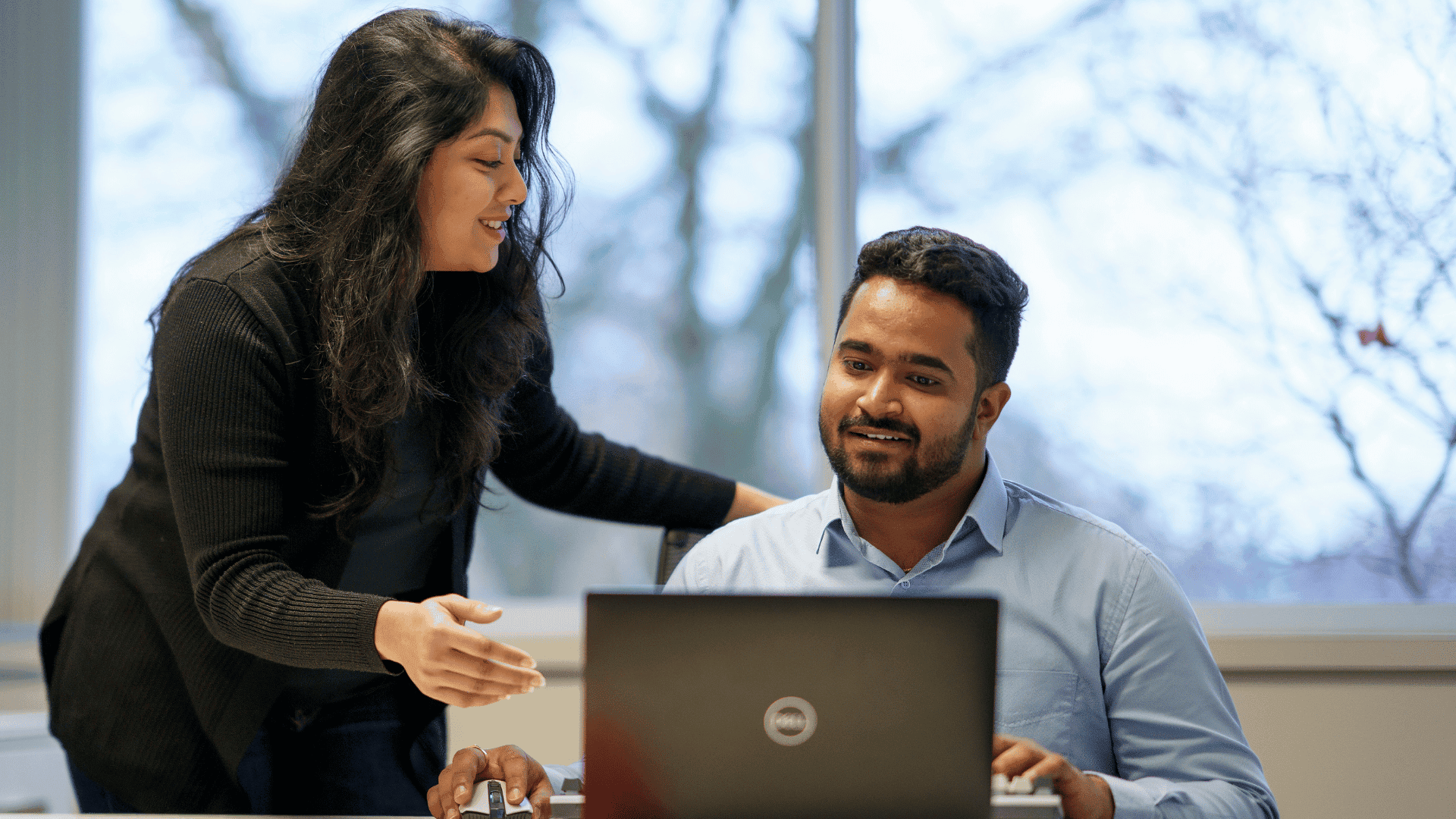 Two people work together at a desk, with one person seated at a laptop and the other standing and gesturing.