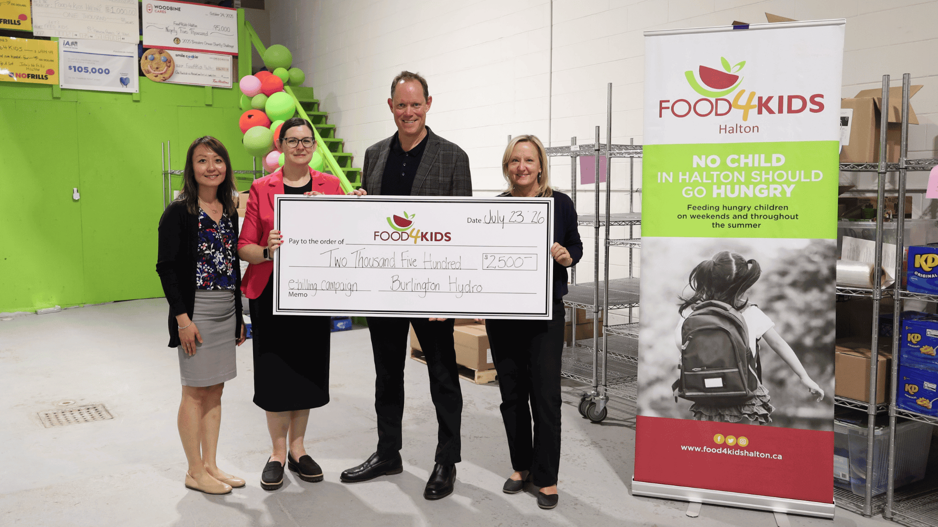 Four people hold a large cheque for $2500 at a Food4Kids Halton donation event, standing next to a banner.