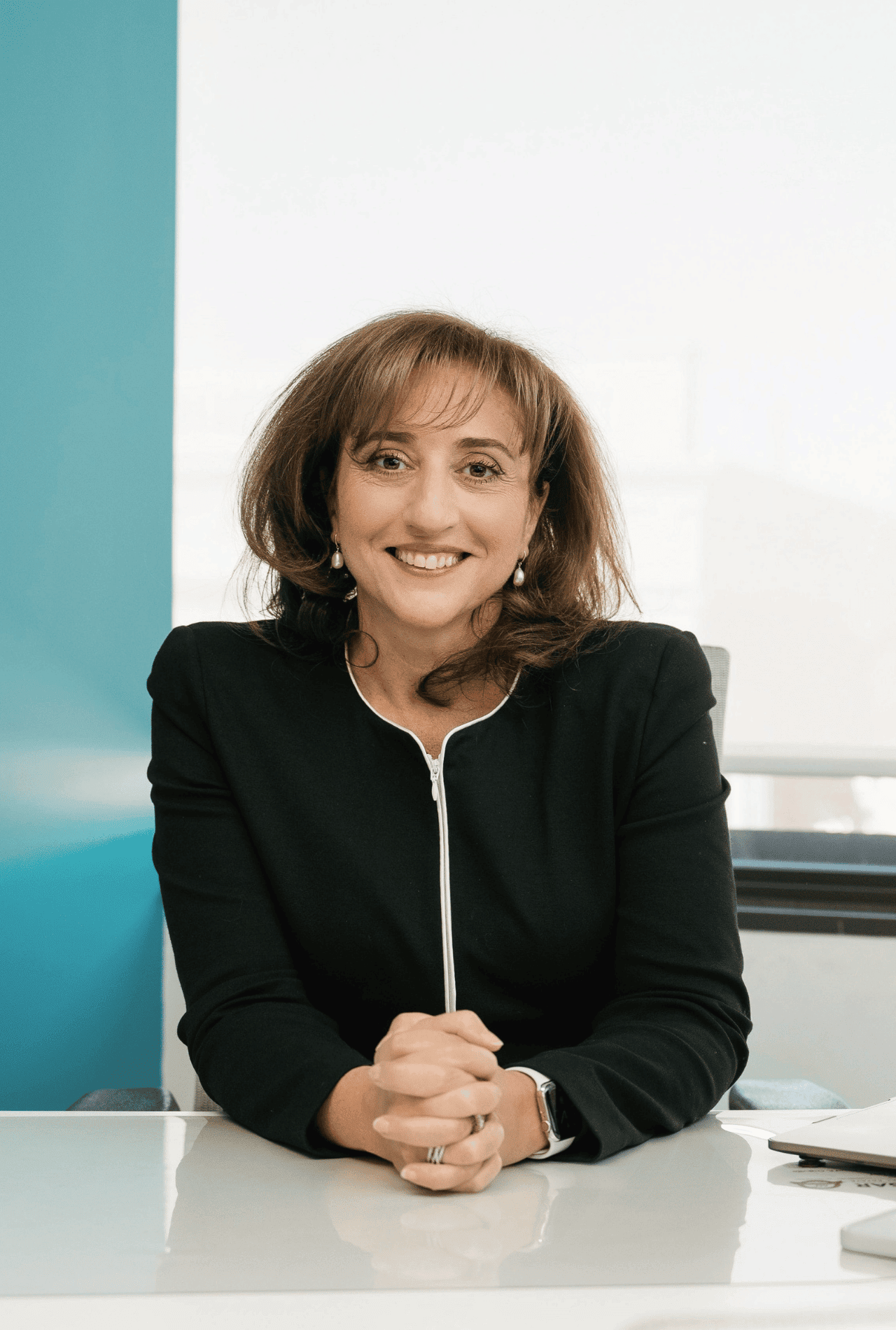Woman in a black jacket sits at a desk, smiling, with hands clasped in front of her in a bright office.